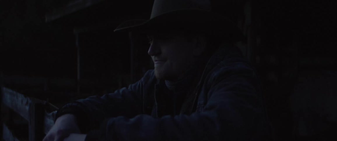A cowboy walks up to a fence and rests his arms on it