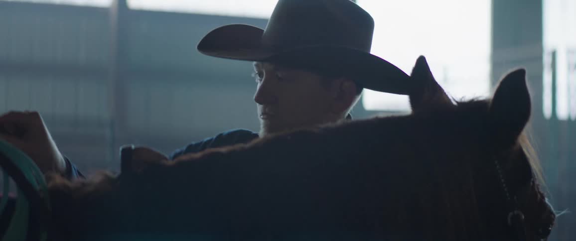A man with a hat securing a saddle on a horse with a wall the sky and clouds in the background