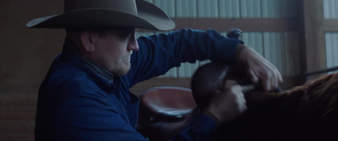 A man with a hat putting on a saddle on a horse with a wall in the background