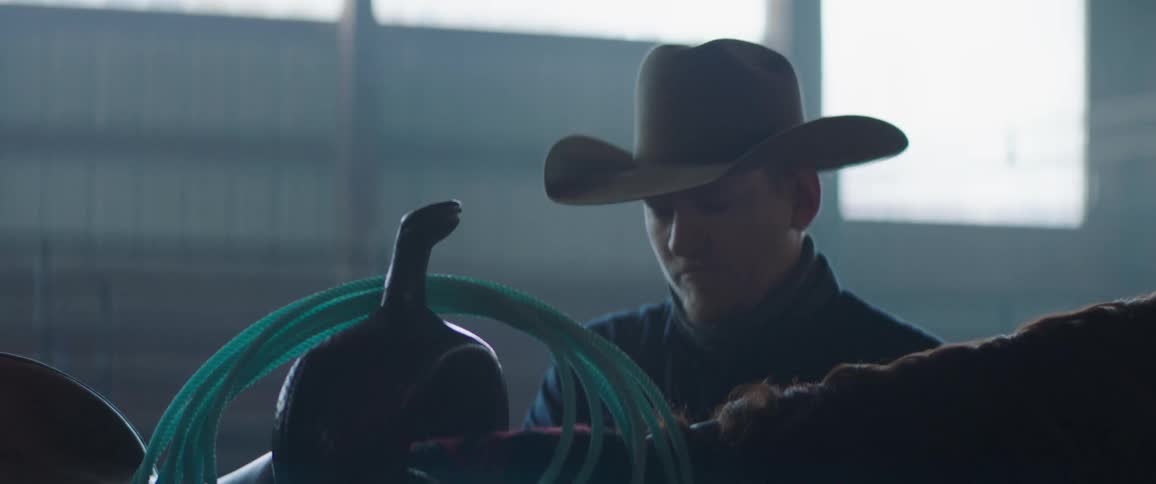 A rancher throws a rope over a saddle on a horse inside of a barn at daytime