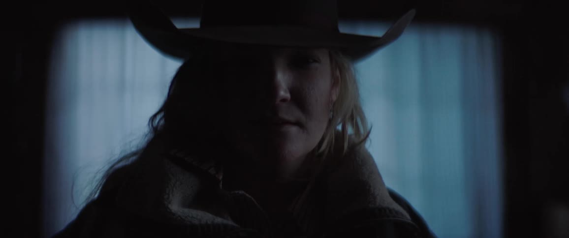 A woman wearing a cowboy hat stands inside of a darkened room while looking directly at the camera