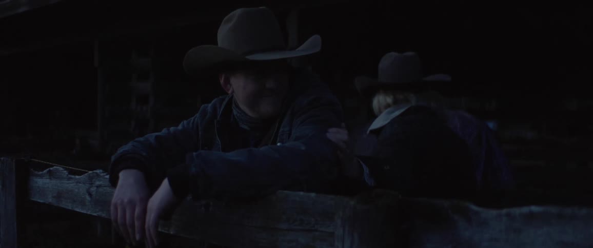 Woman smiles and clasps arm of cowboy leaning against wooden fence