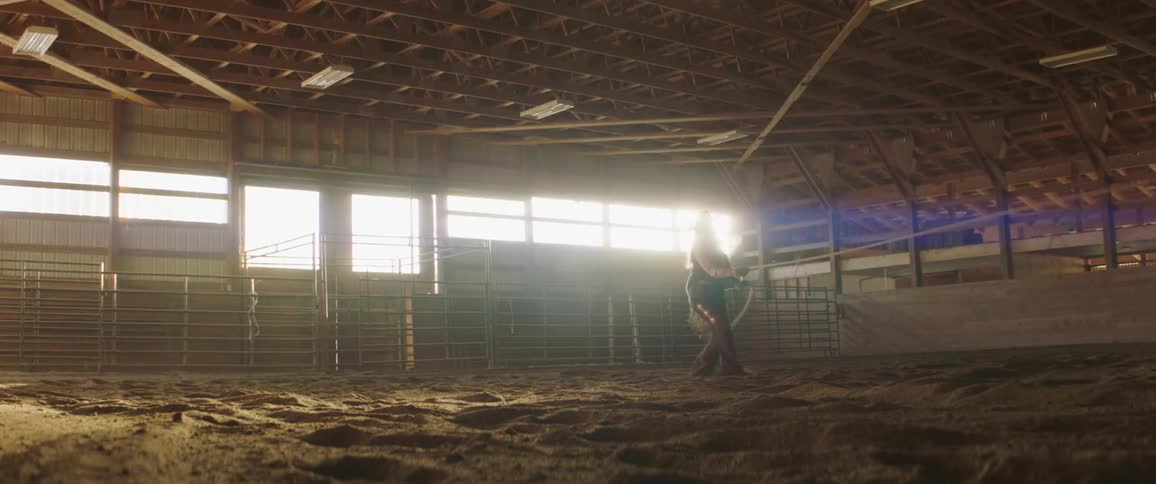 A cowboy holds a rope as a horse runs around a ring while the sun glares through windows