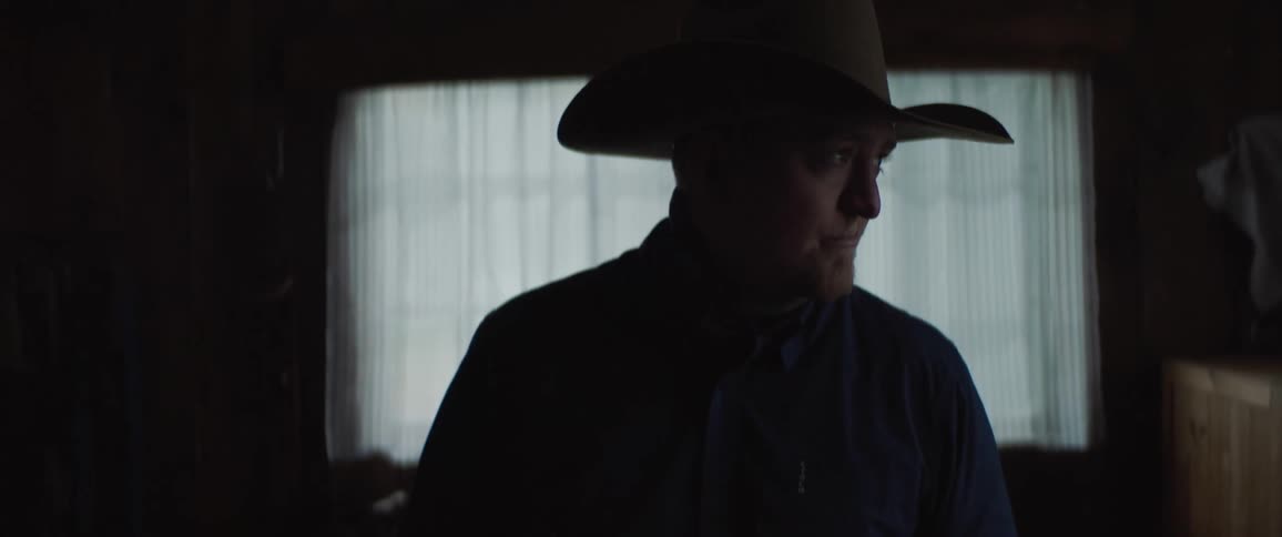 A cowboy stands in a room with a window behind him looking off to the side