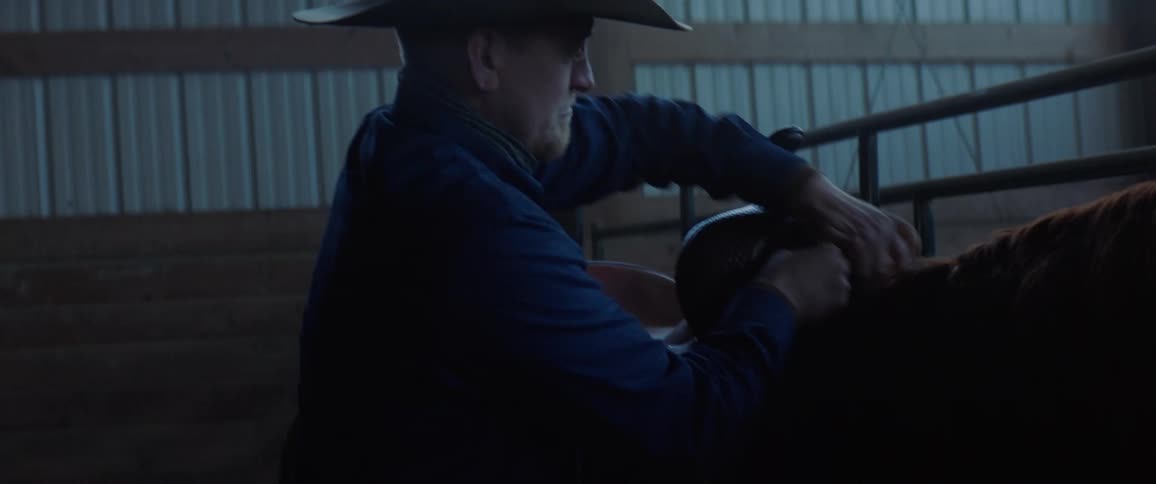 A cowboy fits a saddle onto a horse in a stable or barn