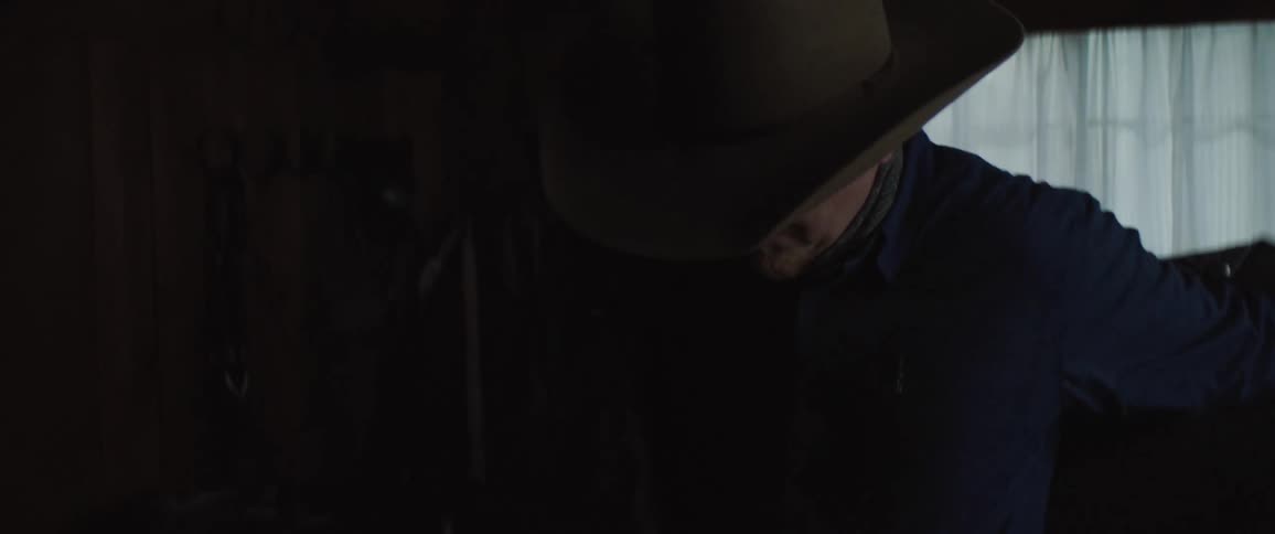 Man in cowboy hat puts on coat in dimly lit room