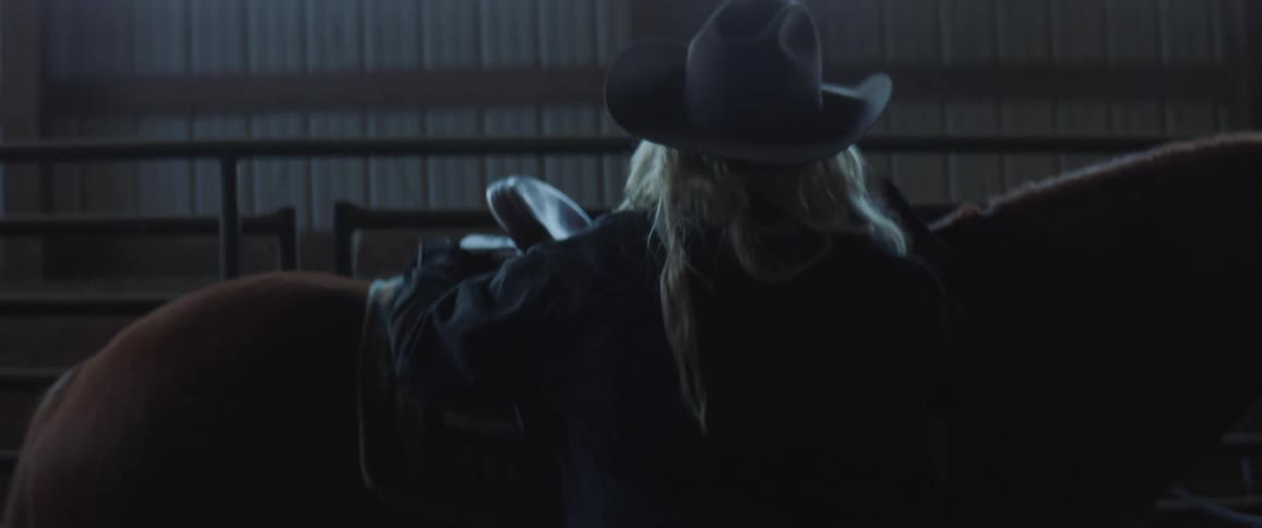 A person with a hat placing a saddle on a horse with a wall and windows in the background