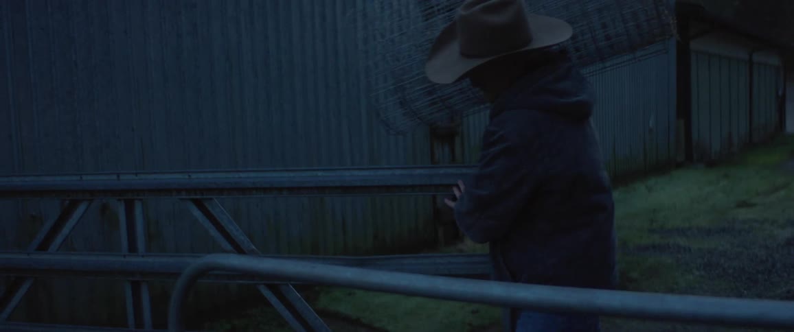 A man with a hat and jacket walking next to a building and opening a fence