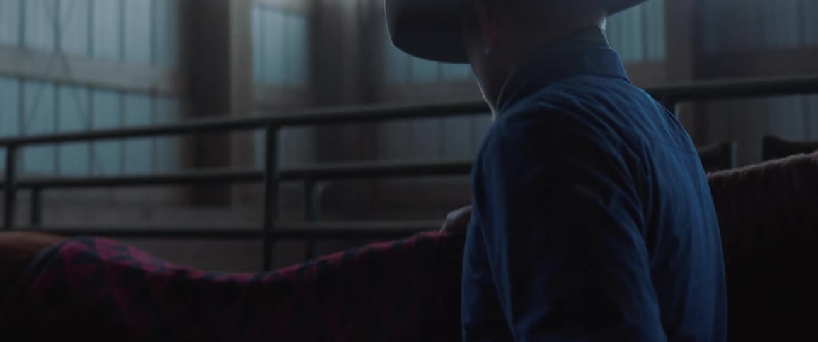A man walks across a stable and puts a saddle on a horse