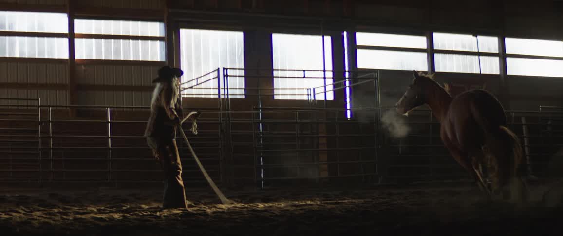 A woman in chaps and cowboy hat swings a rope as a horse runs in a circle around her