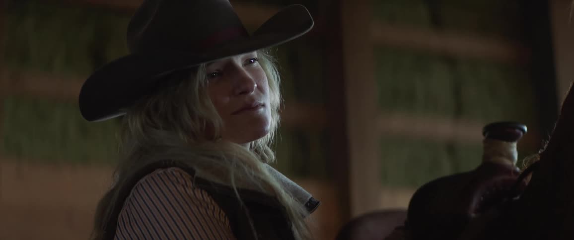 A woman wearing a cowboy hat stands in a barn near a saddled horse looking sad