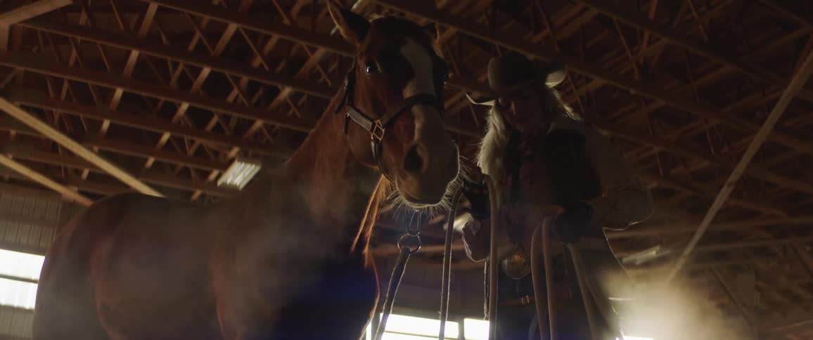 A cowgirl with a rope standing next to a horse in a dark barn