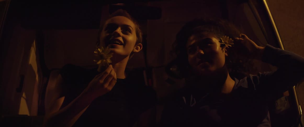 A pair of young women speak while lying next to each other outdoors atop the hood of a car at night