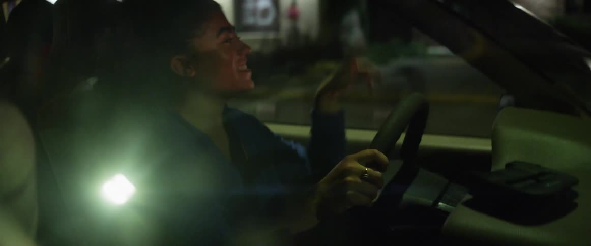 A woman driving a car holding a steering wheel with buildings and hedges in the background