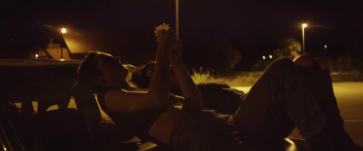 Two women lie on the hood of the their car in a parking lot and play with a flower while looking at the stars