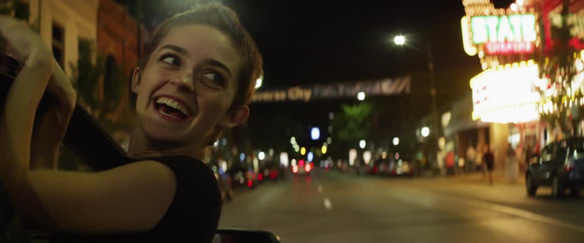 A woman riding in a car with her head out the window with buildings streetlights and trees in the background