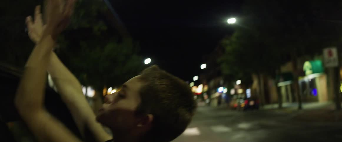 A woman with her head out the window of a moving car with trees and buildings in the background