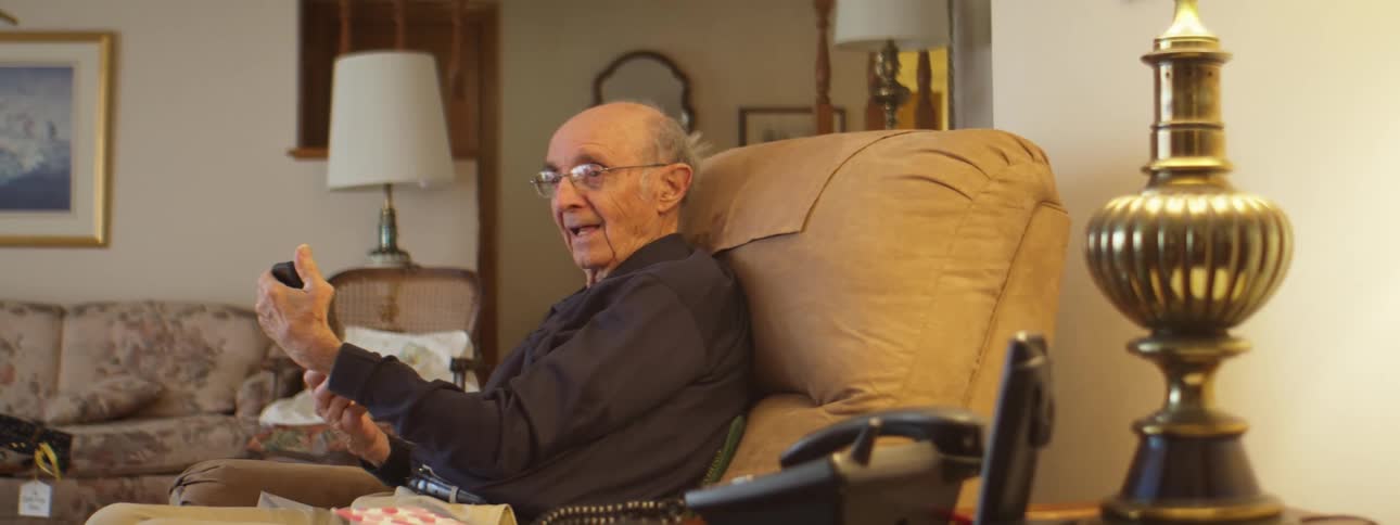 Old bald man with glasses holds a device with shaky hands in the living room at home