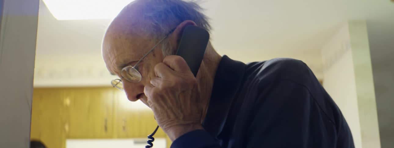 A man in a room picking up a phone and talking with cupboards and a window in the background