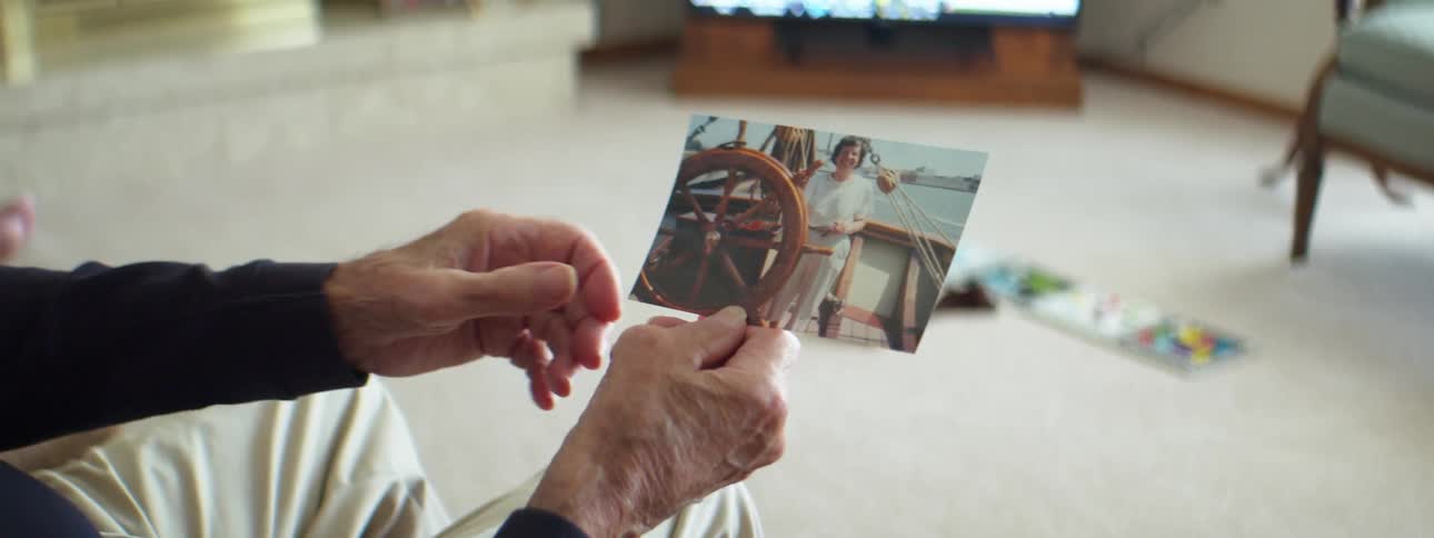 A man sitting indoors on a chair in a living room staring at a photograph hes holding in his hands