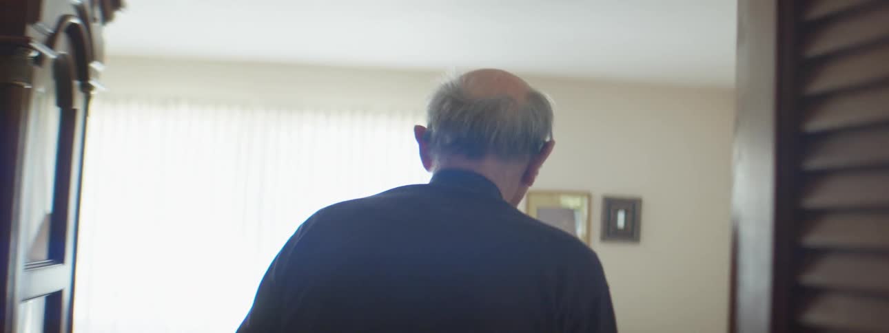 A man walks down a hallway at home into a living room and prepares to sit down