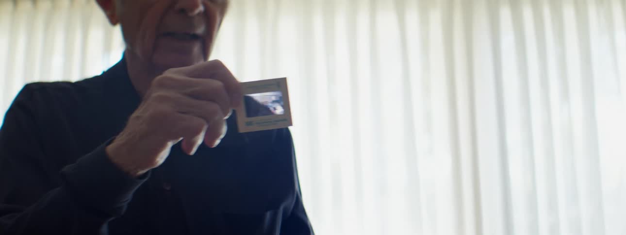 A man in a room holding a projector slide with a window and blinds in the background