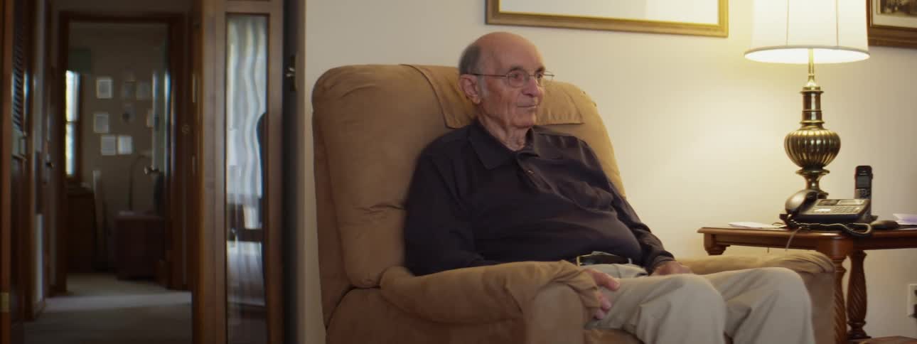 A man with glasses sitting on a couch with a wall and hallway in the background