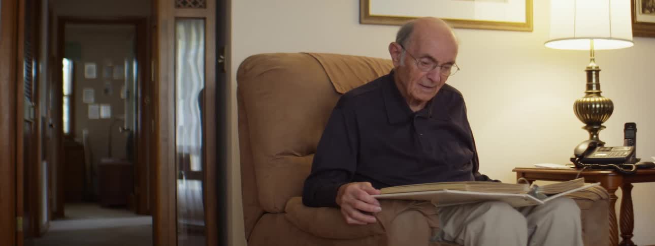 An elderly man sits alone in a chair looking at a large book in his lap