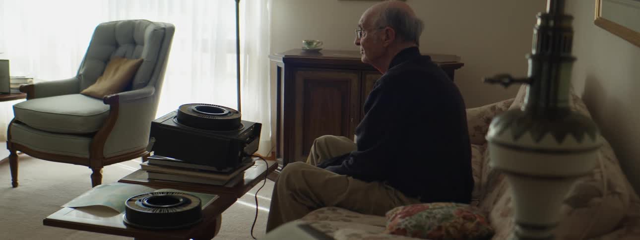 A old man sits on a couch in a living room using an old school projector at daytime