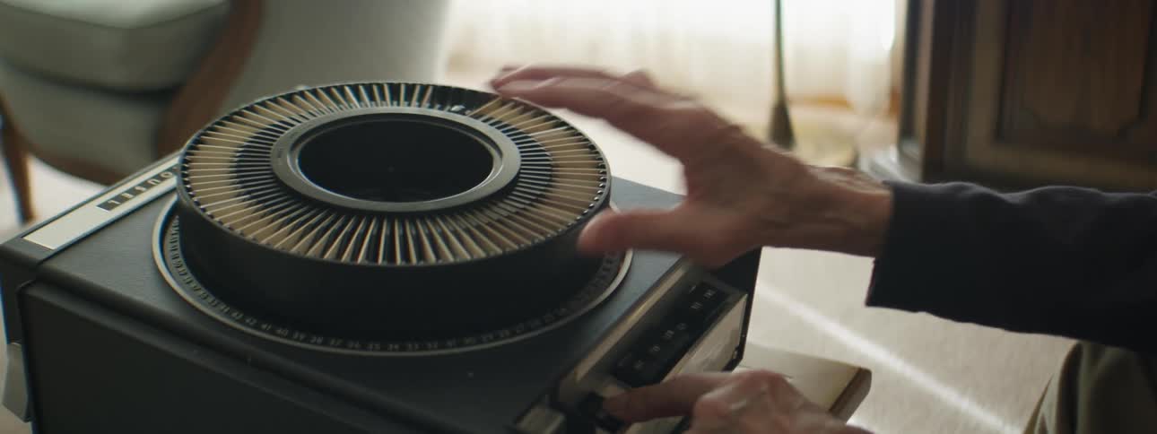 A man adjusting slides on an old school projector while sitting down indoors in a living room at daytime