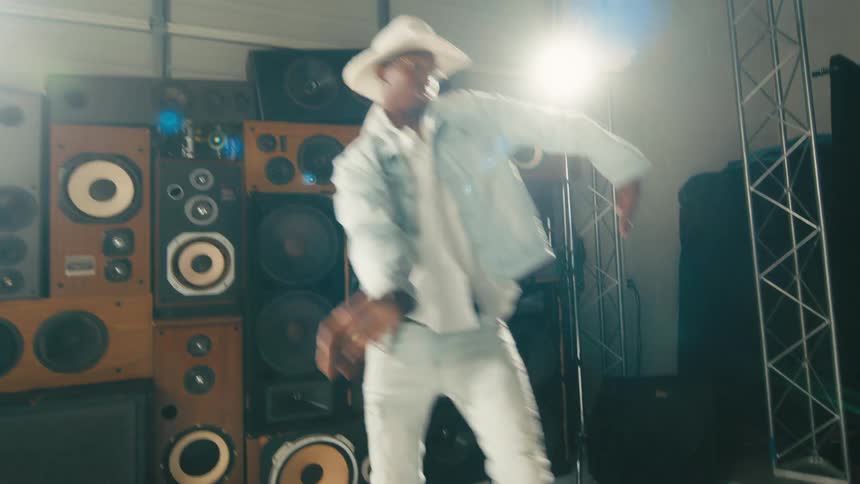 A man with a hat and glasses dancing with a wall and speakers in the background