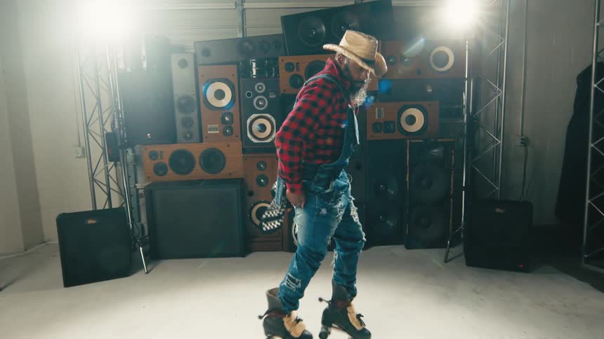 A man with overalls and a hat skating on roller skates with speakers and a wall in the background