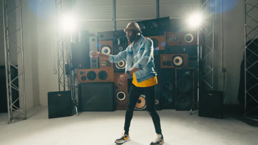 A man dances in a studio in front of old stereos stacked on top of each other next to bright lights