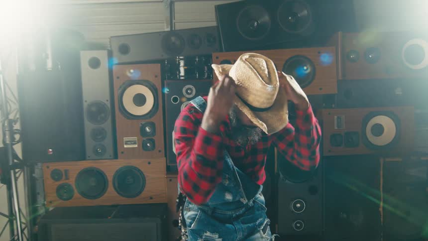 Many speakers line the wall of a studio room with a man dancing on roller skates and wearing a cowboy hat and overalls