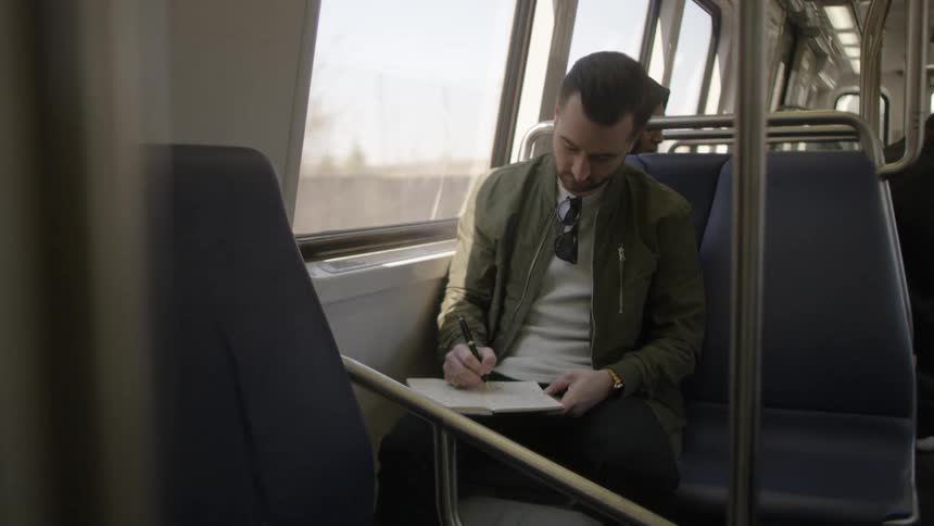 A man writes in a notebook as he rides on a public train through a city
