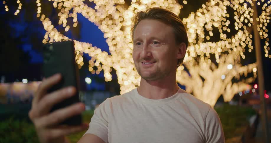 Man in t shirt stands in front of tree decorated with lights conversing via video call on a mobile phone
