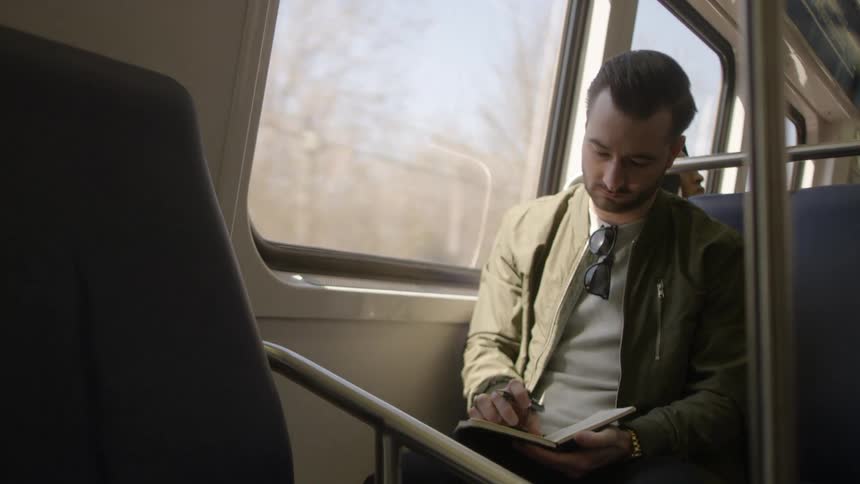 A man riding on a train commuting and writing in a notebook
