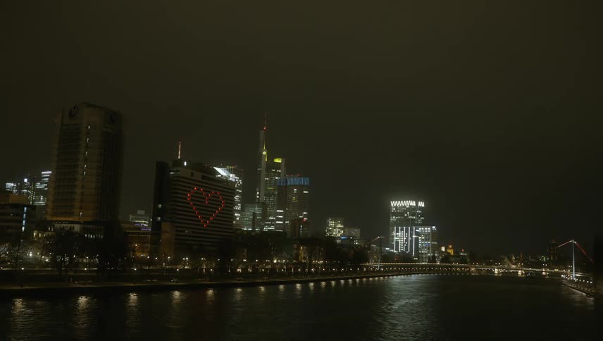 Red lights form a heart shape on the side of a skyscraper that is part of a city skyline along a river