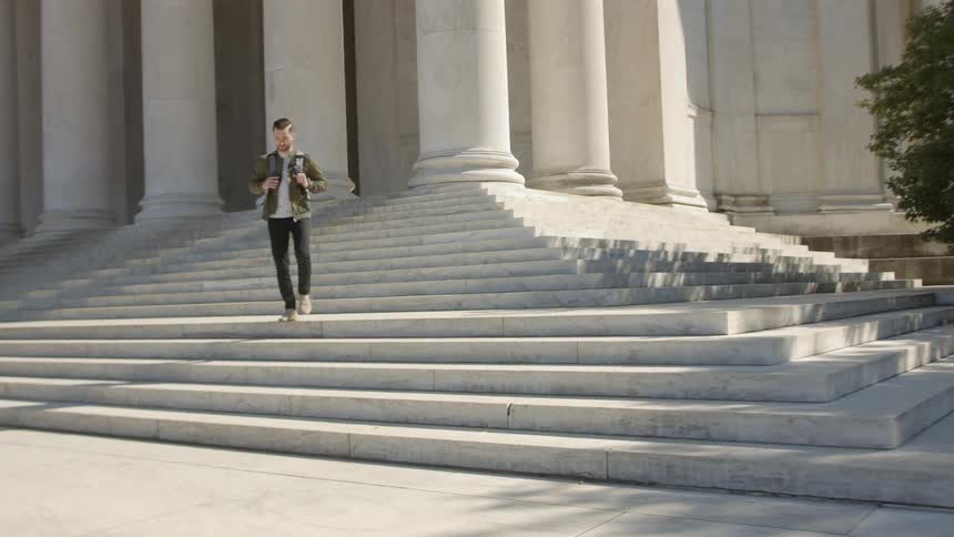 A man holds onto his backpack straps as he walks down the marble steps of a government building with large white columns