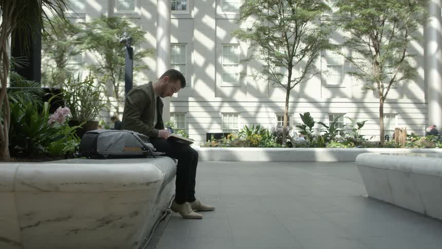 A man sitting down looking at a book with a building plants and trees in the background