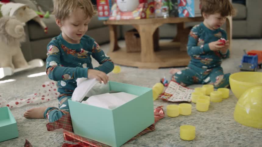 Two boys with pajamas on the floor with one of the boys opening a present with a table and a sofa in the background