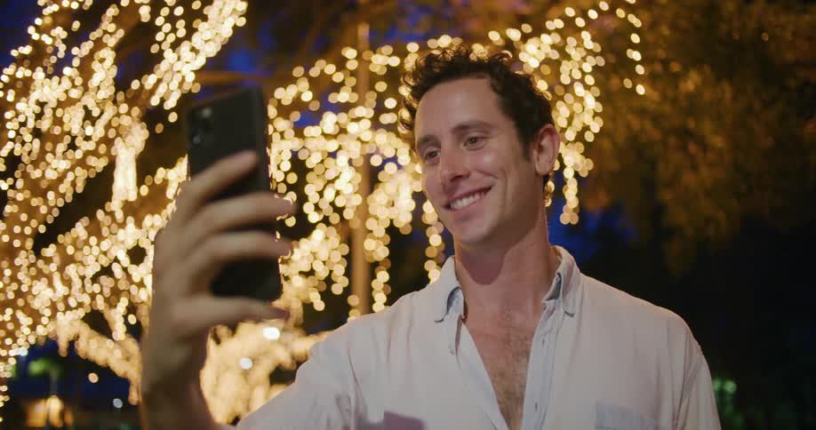 A man holding up his phone during a video call blowing kisses and laughing with a lighted tree behind him