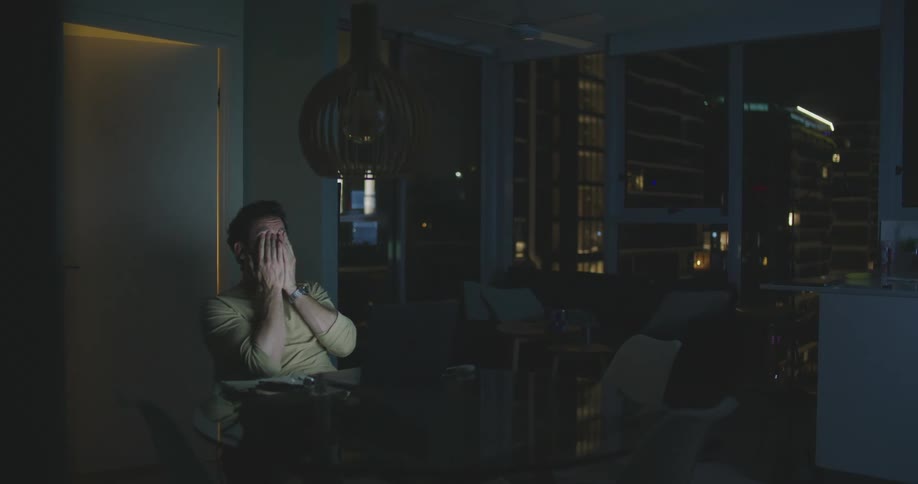 A man sits at his desk in an office at night and types on his laptop and then rubs his face with his hands