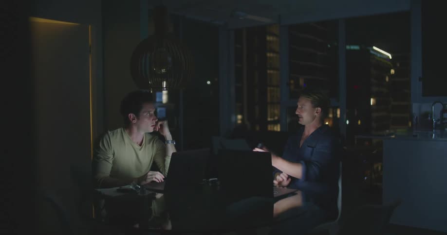 Two men sitting by a table in front of laptops with a door and window in the background