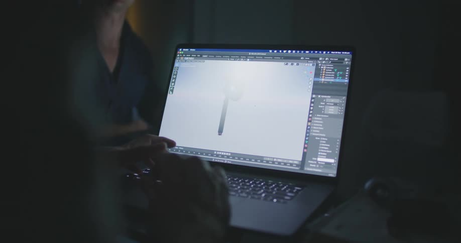 A man points at a computer screen while a person makes adjustments to a design in a computer software program indoors at nighttime
