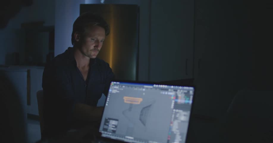 A man sits behind another person who is on a laptop using graphic designing software in a living room at nighttime