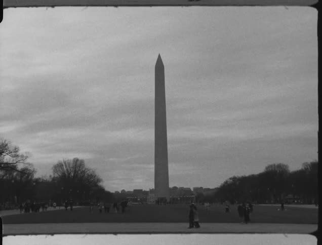 The washington monument looms over the pedestrians walking along the path and field below