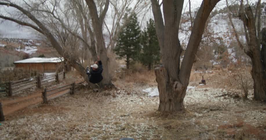 Man jumps off slope onto rope swing at farm with snow covered hill in background