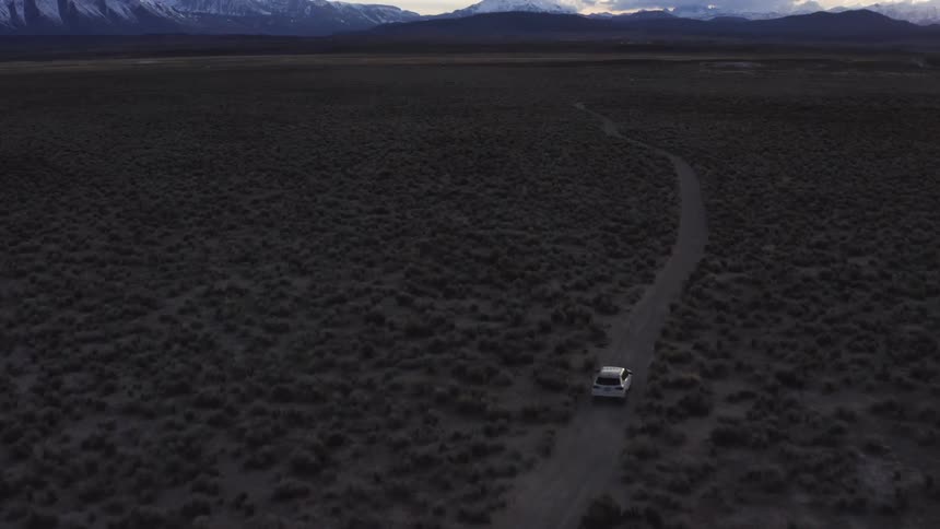 A vehicle driving on a dirt road in the desert with a mountain range the sky and clouds in the background