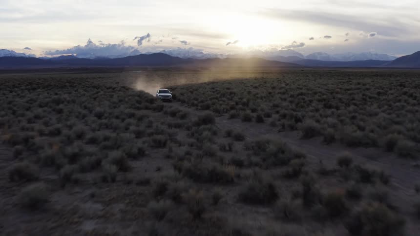 Suv drives along dirt road as sun shines from behind clouds over mountains